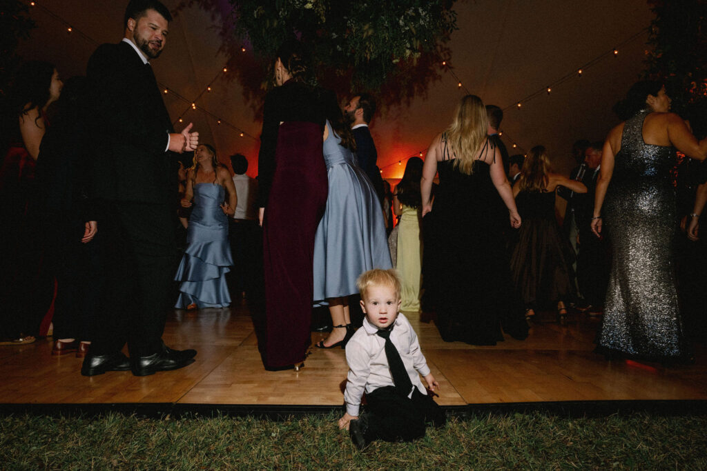 A young child sits on the edge of the dance floor while guests dance inside a tented wedding reception in Lake Placid.