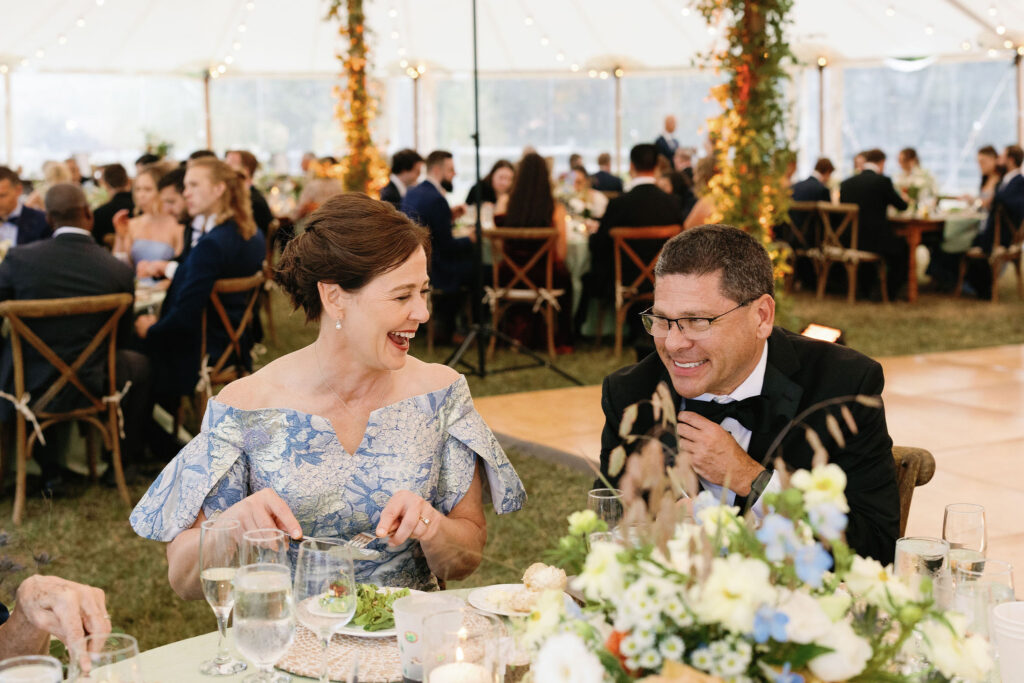 Guests laugh together over dinner with floral centerpieces inside a sailcloth tent at a Lake Placid, New York wedding.