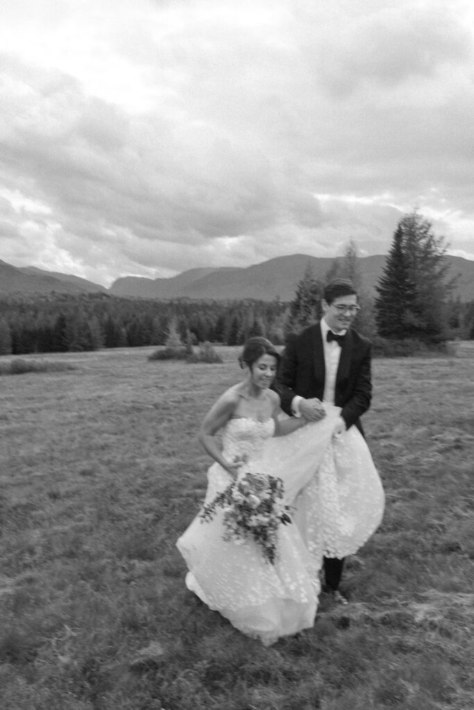 Bride and groom walk and laugh together across an open field with Adirondack mountains in Lake Placid, New York.