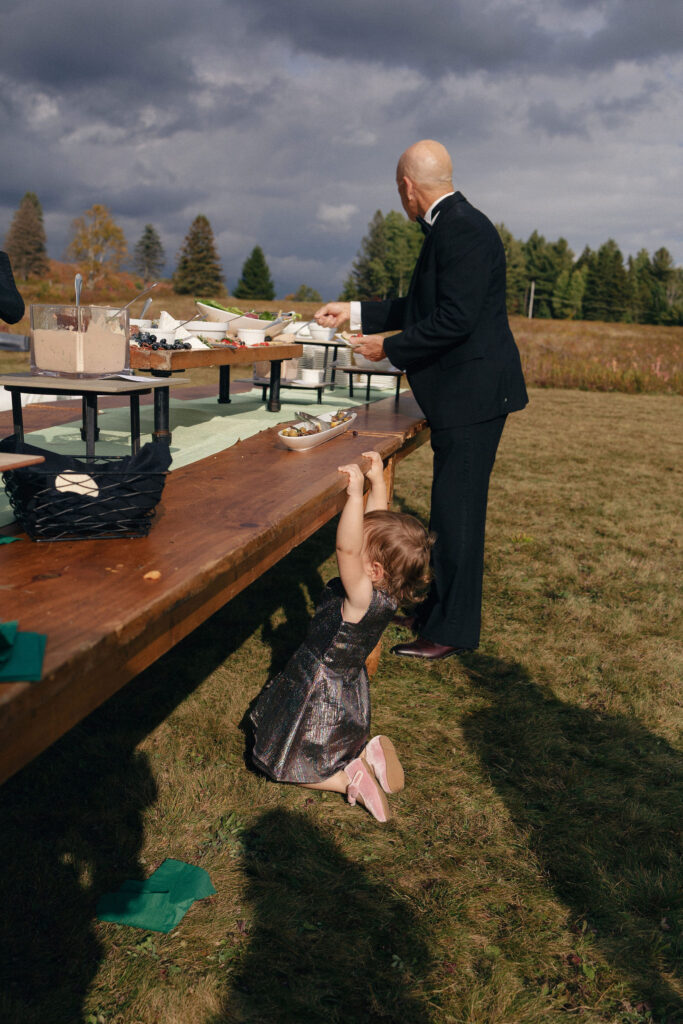 A guest serves food at an outdoor buffet while a young child reaches up to the table during a Lake Placid wedding.