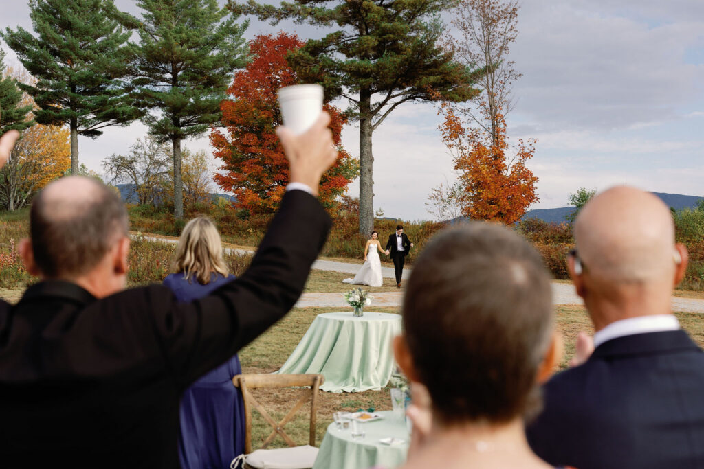 Guests raise drinks as the bride and groom walk together across a field with fall foliage during a Lake Placid wedding.