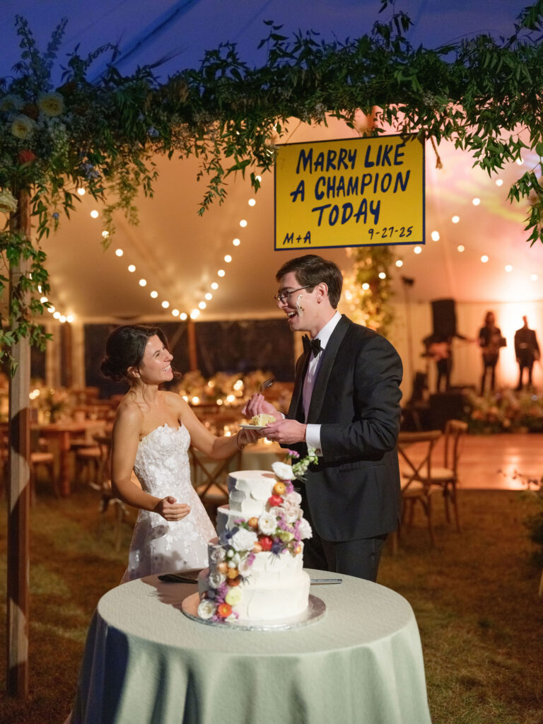 Bride and groom laugh while cutting their wedding cake beneath string lights in a tented Lake Placid reception.