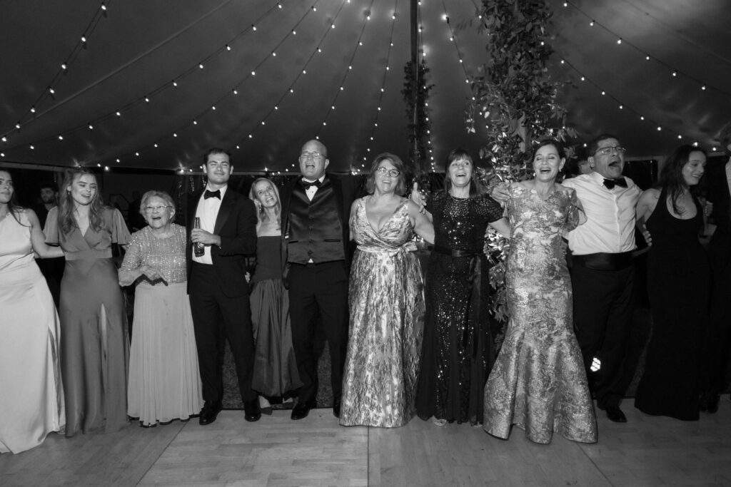 Guests stand arm in arm singing and dancing together under string lights inside a Lake Placid wedding tent.