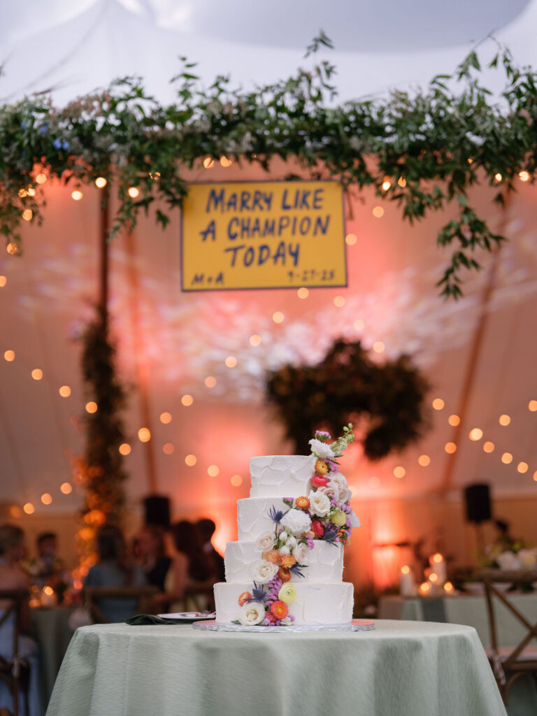 Three-tier wedding cake with fresh flowers displayed beneath a “Marry Like a Champion Today” sign at a Lake Placid reception.