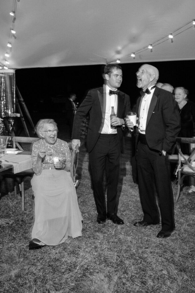 Wedding guests in formal attire laugh together beneath string lights inside a tented reception in Lake Placid, New York.