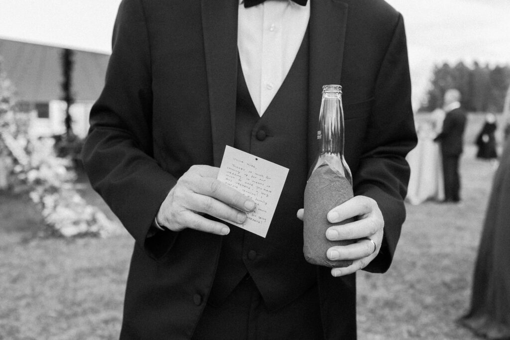 Groom in a tuxedo holds a handwritten note and a drink during cocktail hour at a Lake Placid wedding.