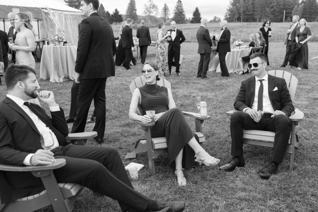 Wedding guests relax in Adirondack chairs with drinks during cocktail hour at a Lake Placid, New York outdoor wedding.