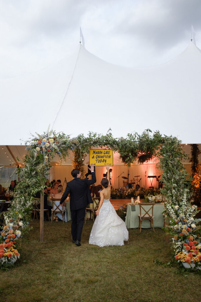 Bride and groom enter the reception under a floral-covered arch with a “Marry Like a Champion Today” sign at a Lake Placid wedding.
