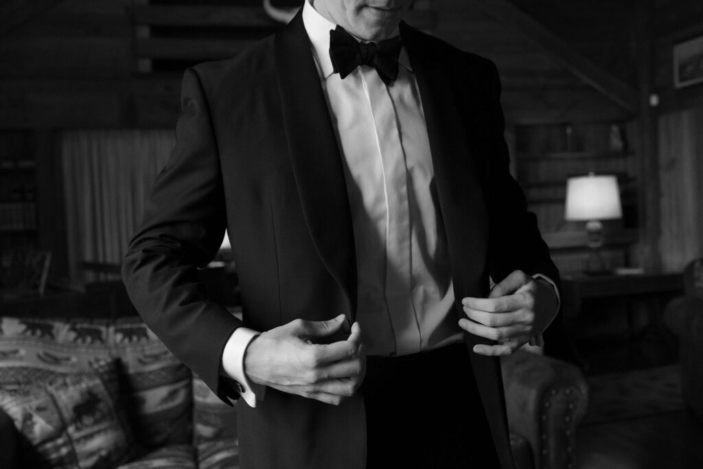Groom adjusting tuxedo jacket inside rustic cabin at River Ranch Lake Placid wedding