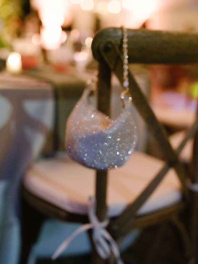 Beaded clutch purse hangs from the back of a wooden reception chair with candlelit tables softly blurred in the background at a Lake Placid wedding.