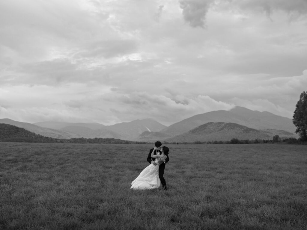 Bride and groom embrace in a wide Adirondack field with dramatic mountains and clouds in Lake Placid, New York.