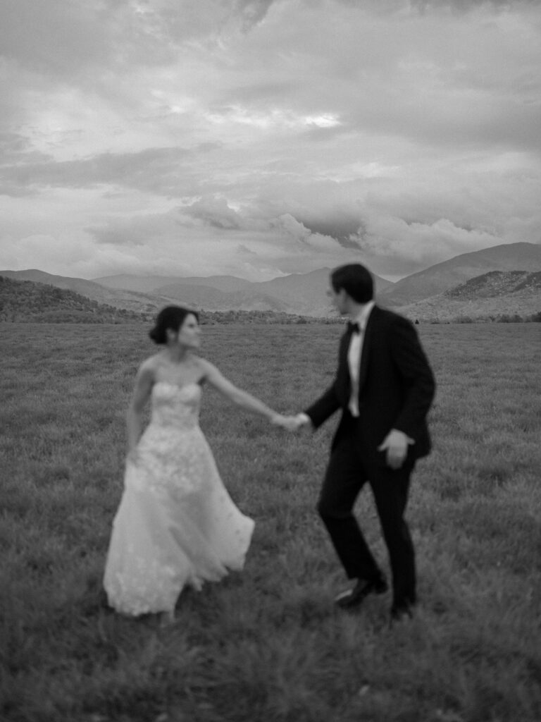Bride and groom hold hands while walking through an Adirondack field with mountains in Lake Placid, New York.