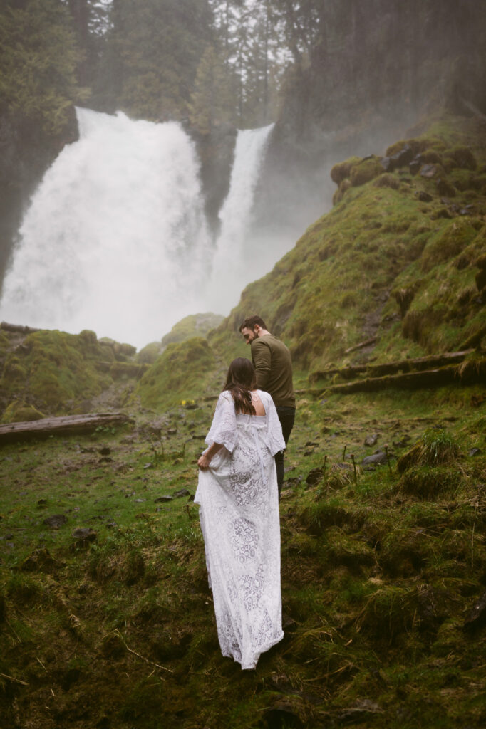 A bride and groom walk hand-in-hand away from the camera towards Sahalie Falls in Oregon. The groom is slightly ahead of the bride and is looking back at her as they walk.