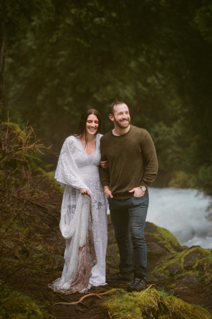 A bride and groom stand at the base of Sahalie Falls in Oregon. The groom has his hands in his pockets while the bride stands next to him, holding his arm. Neither look at the camera as they are laughing.