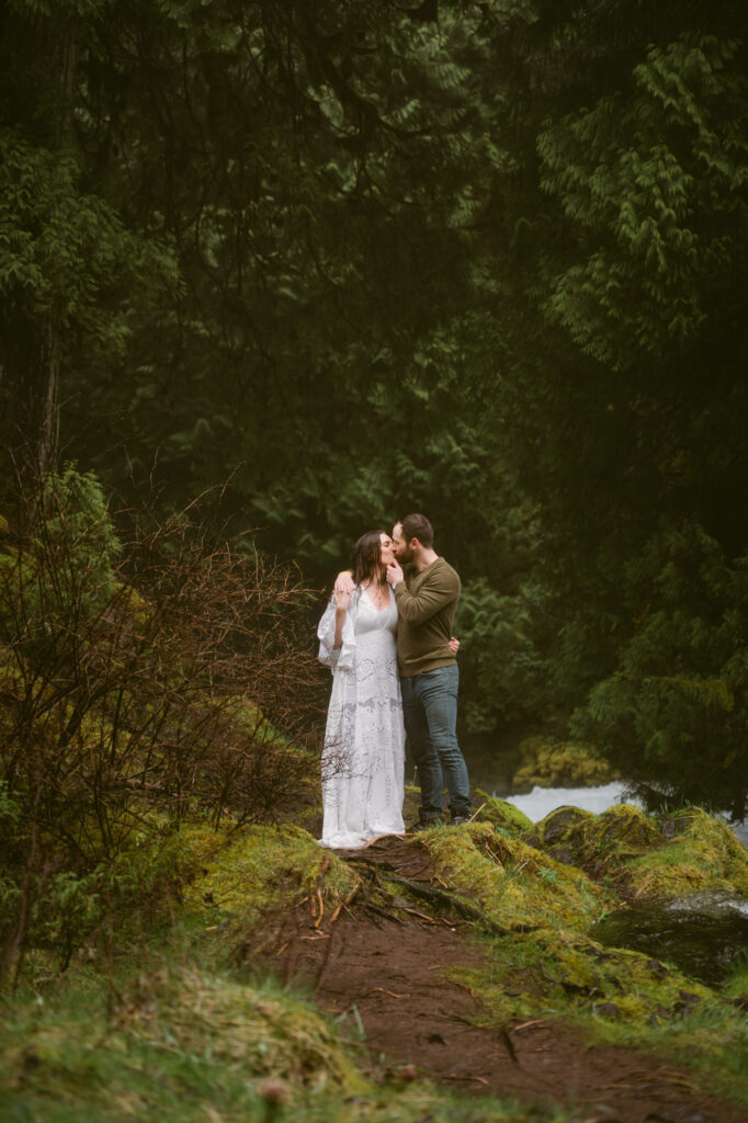 A bride and groom stand at the base of Sahalie Falls in Oregon. The bride has one arm around the groom's waist, pulling him close. The groom is using one hand to lift her chin, kissing her on the lips.