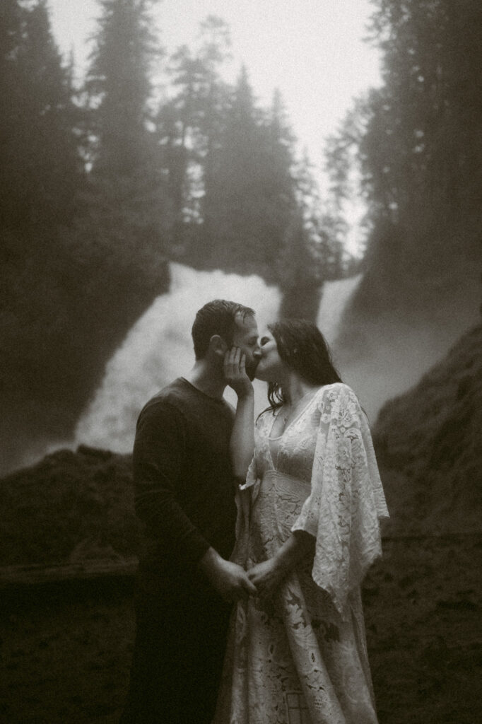 A black and white image of a bride and groom standing at the base of Sahalie Falls in Oregon. The bride is pressed against the groom's chest so they form a T with their bodies. She's using her inside arm to cup the groom's cheek as they kiss.