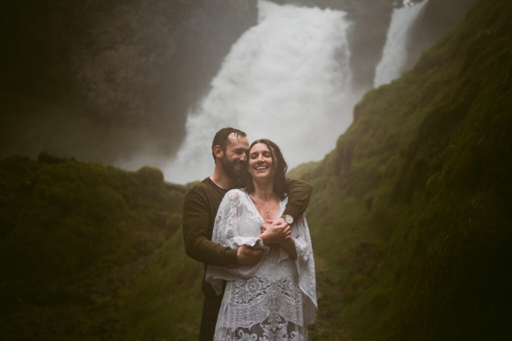 A bride and groom stand at the base of Sahalie Falls in Oregon. The groom is standing behind the bride with his arms wrapped around her while the bride holds his hands. Both are laughing as they get soaked with the spray coming off of the falls.