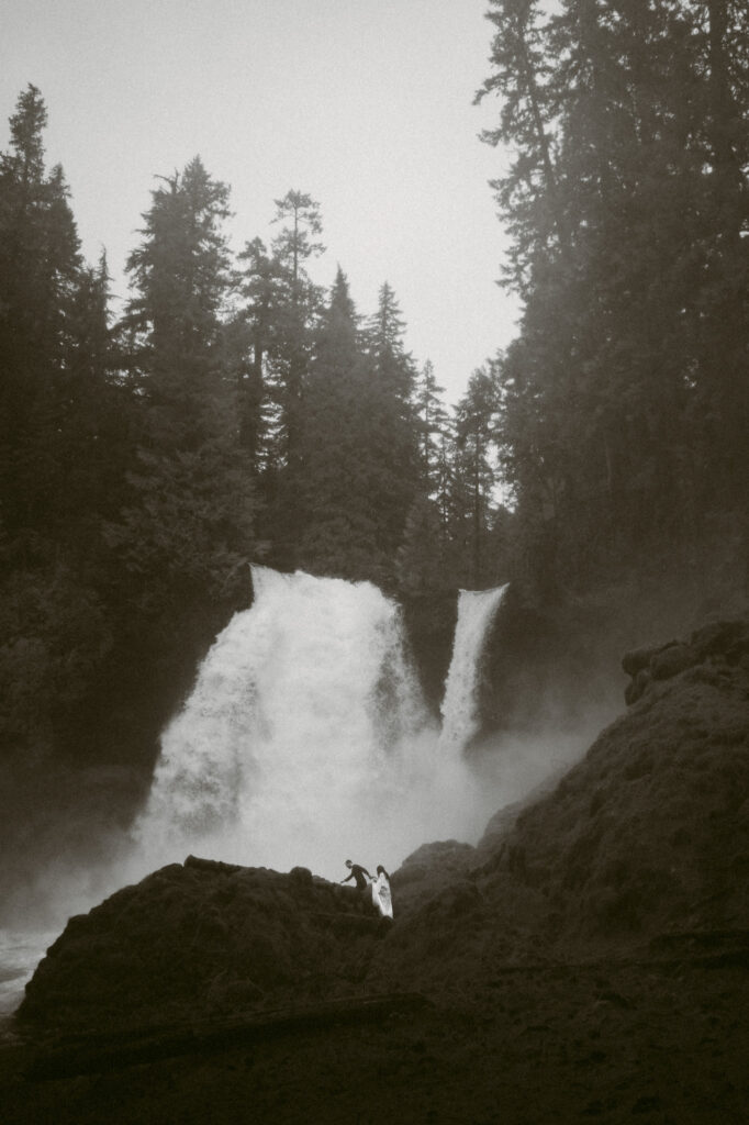 A black and white pulled back landscape shot showing Sahalie falls with a small bride and groom walking towards it.