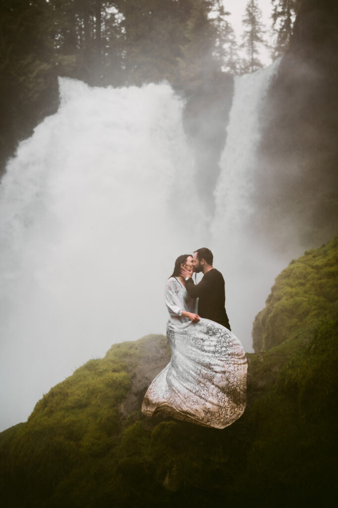 A bride and groom stand at the base of Sahalie Falls, getting soaked by the spray. The groom is holding the bride's face and kissing her as she swings the train of her muddy wedding dress in front of them both.
