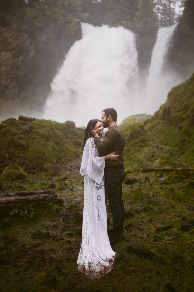 A bride and groom stand at the base of Sahalie Falls in Oregon. The bride has her hands on the groom's waist while the groom is holding her face, kissing her temple.
