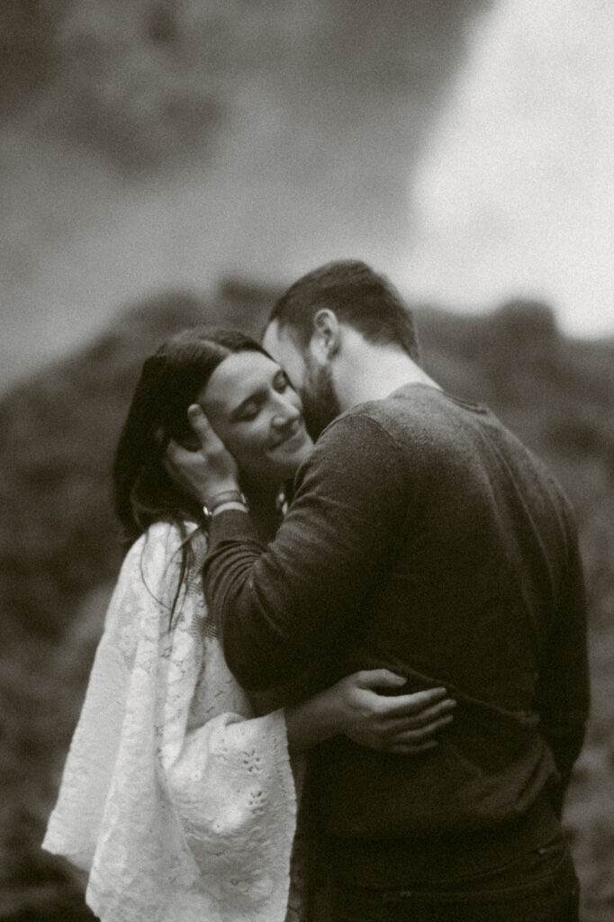 A black and white image of a bride and groom standing at the base of Sahalie Falls in Oregon. The bride has her hands on the groom's waist while the groom is holding her face, kissing her temple.