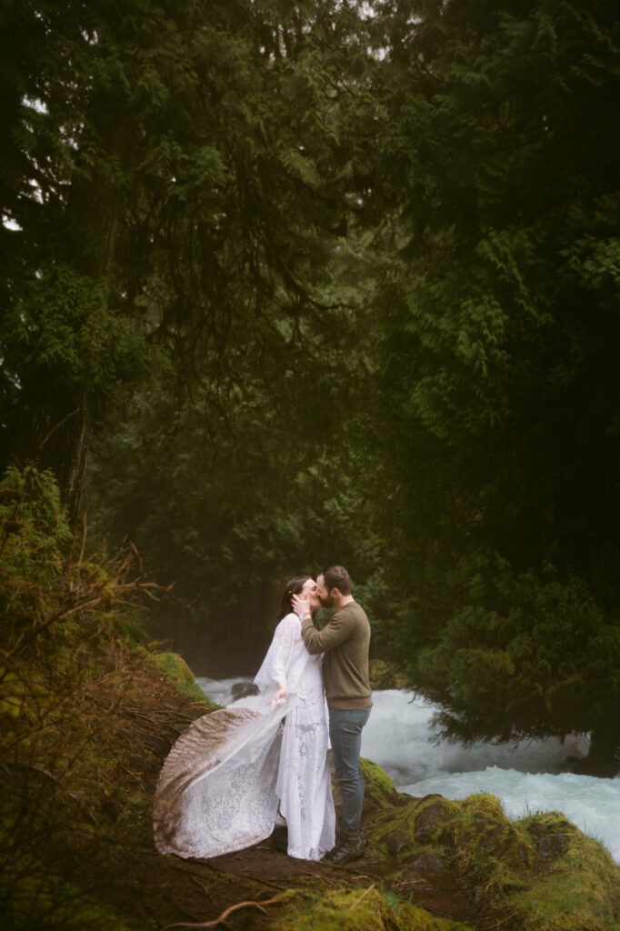 A bride and groom stand at the base of Sahalie Falls in Oregon. The groom holds the bride's face, kissing her, as the bride swings the train of her dress back and forth.