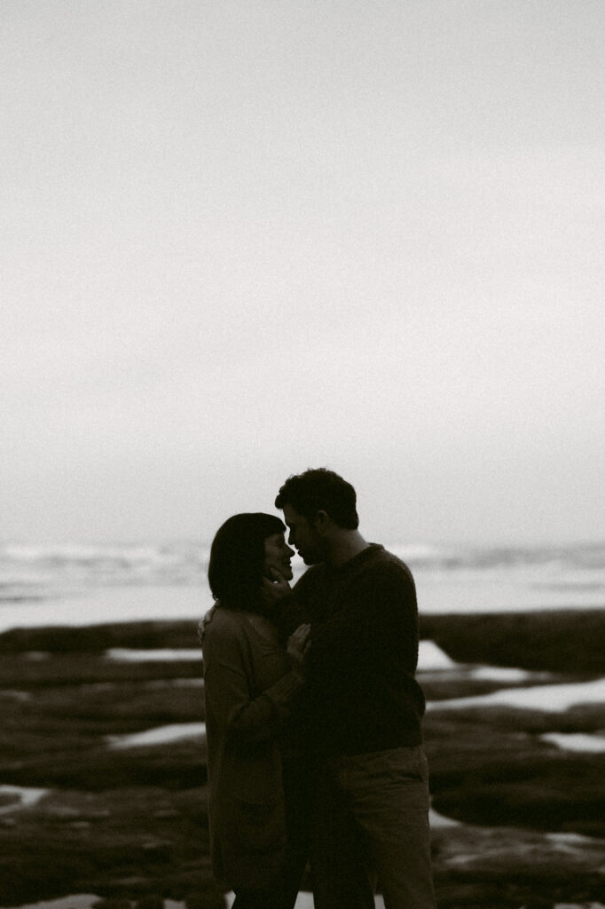 A black and white image of a man and woman silhouetted against the pacific ocean, about to kiss. 