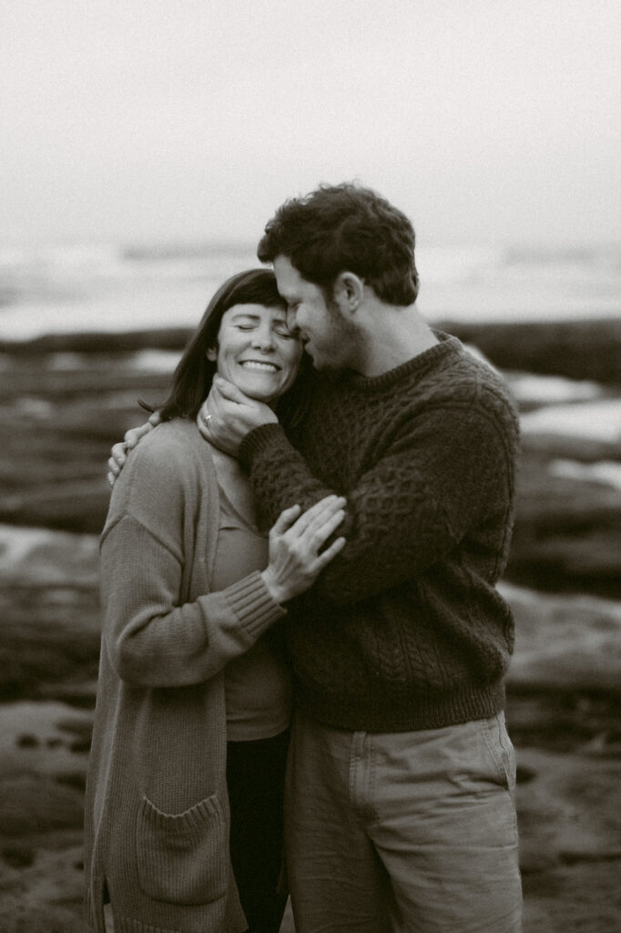 A black and white image of a man and woman in front of the pacific ocean at Otter Crest beach in Oregon. The man has one hand on the woman's back, the other under her chin. The woman has her hand on the man's elbow and is leaning against him, smiling, as he kisses her cheek.