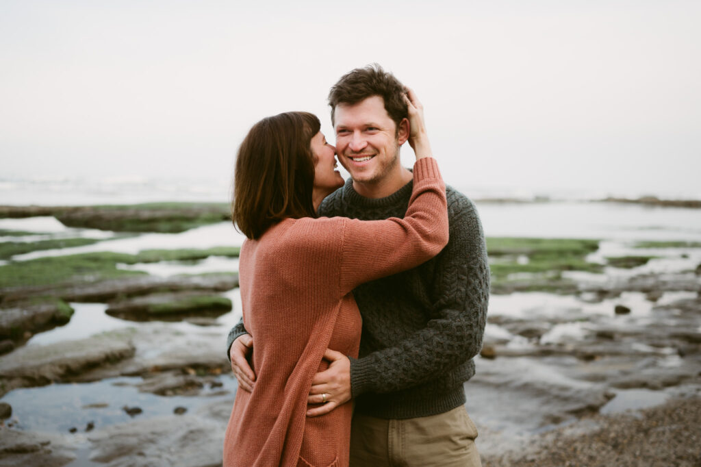 A man and woman stand in front of the pacific ocean at Otter Crest beach. The man's hands are on the woman's waist and she is touching his head, about to kiss his cheek.