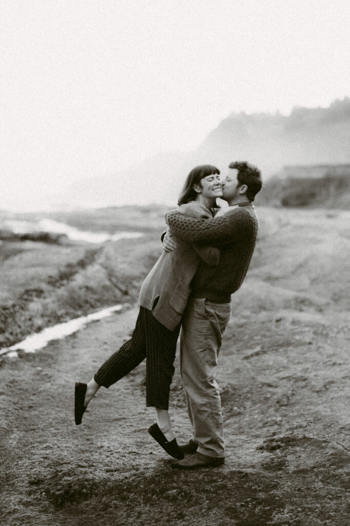 A black and white image of a man and woman standing in front of Devil's Punchbowl in Oregon. The man is lifting the woman off her feet and kissing her cheek. The woman is smiling.