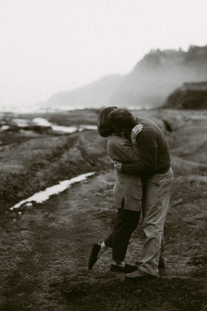 A black and white image of a man and woman by Devil's Punchbowl in Oregon. The man has his hands on the woman's waist and is leaning her back while kissing her neck.