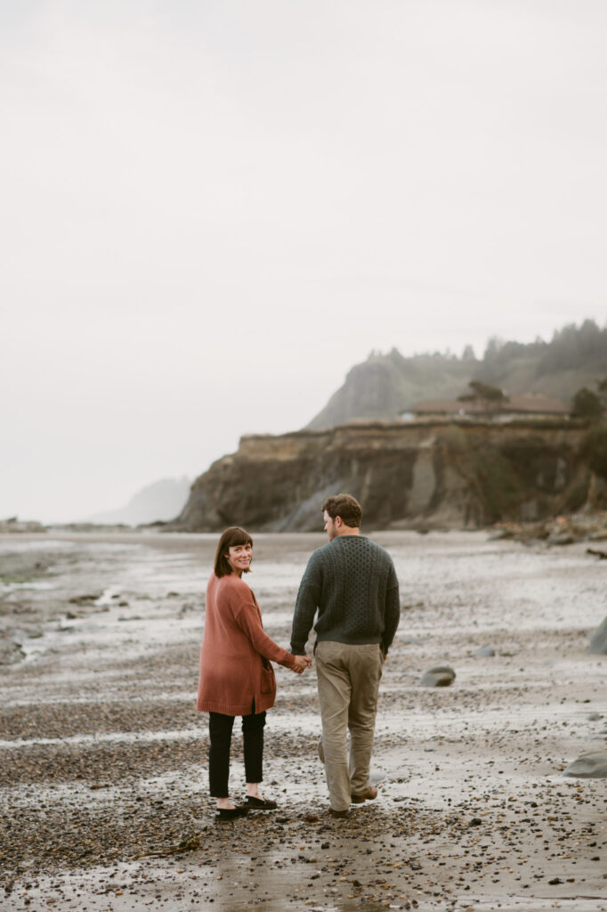 A man and woman walk hand in hand on the shoreline of Otter Crest Beach. They're walking away from the camera with the woman glancing over her shoulder at it.