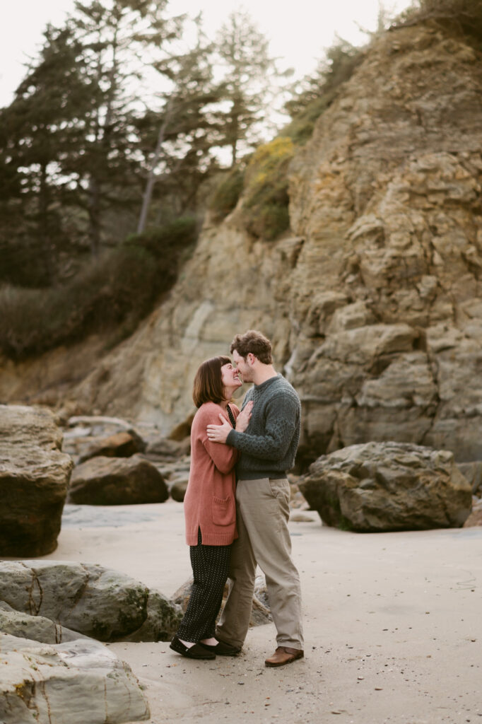 A man and woman stand amongst the rocks on the shoreline of Otter Crest Beach in Oregon. The woman's hands are on the man's chest and they are leaning towards one another to kiss.