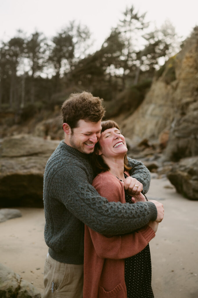 A man and woman stand on Otter Crest Beach in Oregon. The man's arms are wrapped around the woman and her back is against his chest. They are holding hands and laughing.