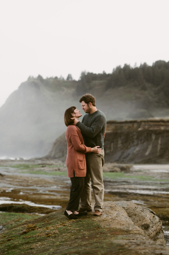 A man and woman standing on the rocks at Otter Crest Beach in Oregon. There's a mist behind them as the man gently cups the woman's face, tilting her up to face him.