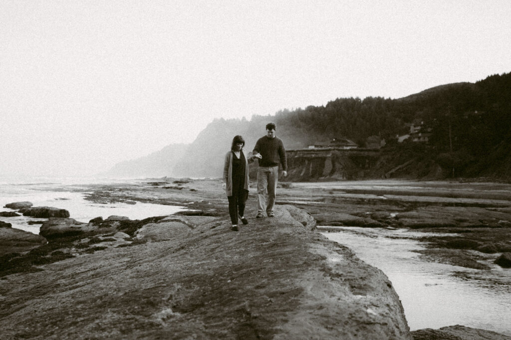 A black and white image of a man and woman walking along the rocky shoreline at Devil's Punchbowl in Oregon.