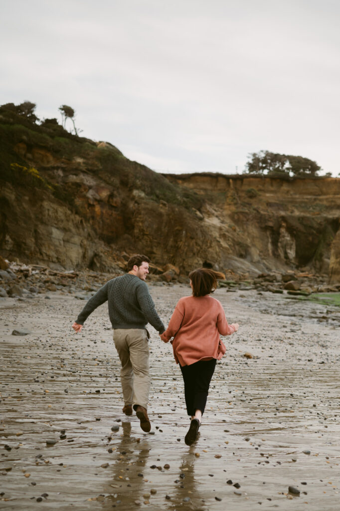 A man and woman hold hands as they run down Otter Crest beach in Oregon. They are looking at one another and running away from the camera.