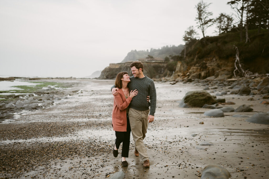 A man and woman stroll down the shore of Otter Crest Beach in Oregon. The man's arm is around the woman's shoulders and she is grasping onto his waist. They're looking at each other and smiling.
