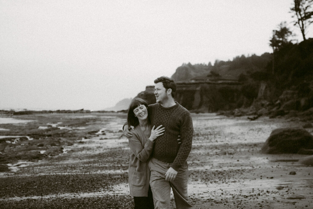 A black and white image of a man and woman walking down the shoreline of Otter Crest beach in Oregon. The man's arm is around the woman's shoulders and she is laughing at the camera while he looks out to sea.