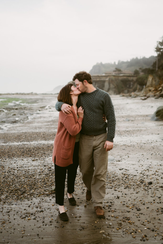 A man and woman walk down the coastline of Otter Crest beach in Oregon. The man's arm is around the woman's shoulders and he is pulling her in for a kiss.
