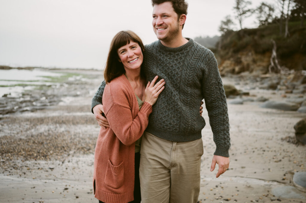 A man and woman walk down the shoreline of Otter Crest Beach in Oregon. The man's arm is around the woman's shoulders. She's smiling at the camera while he looks out to sea, also smiling.