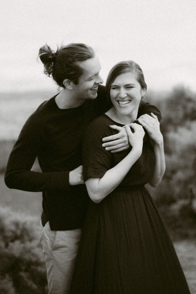 A black and white image of a man and woman standing close together. The man's arm is around the woman's chest while the woman holds onto it with both hands. They are leaning into one another, laughing.