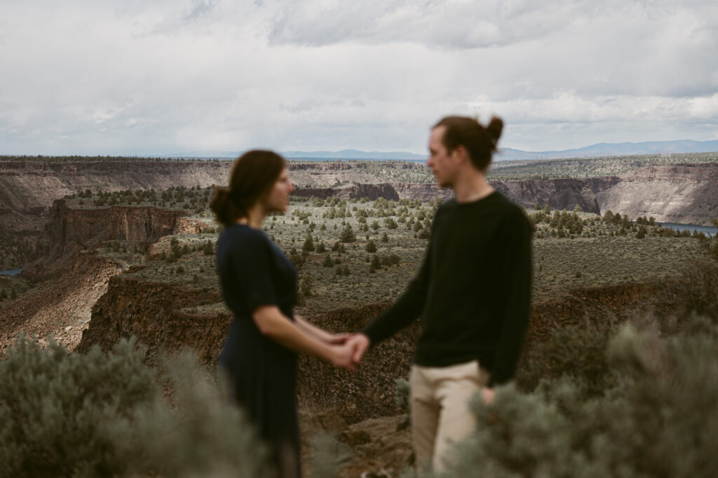 An out of focus shot of a man and woman holding hands, the background is in focus showing Cove Palisades state park.