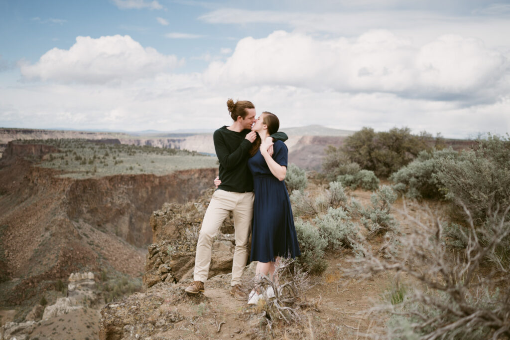 A man and woman stand at the edge of the Tam-a-lau trail at Cove Palisades State Park. The man has one arm around the woman's shoulders, the other is under her chin, lifting her face up for a kiss.