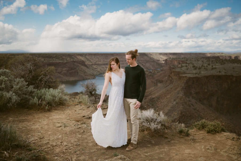 A man and woman stand at the Tam-a-lau Trail overlook at Cove Palisades State Park in Oregon. The woman is wearing a white dress and holding it in front of her so it catches the breeze.
