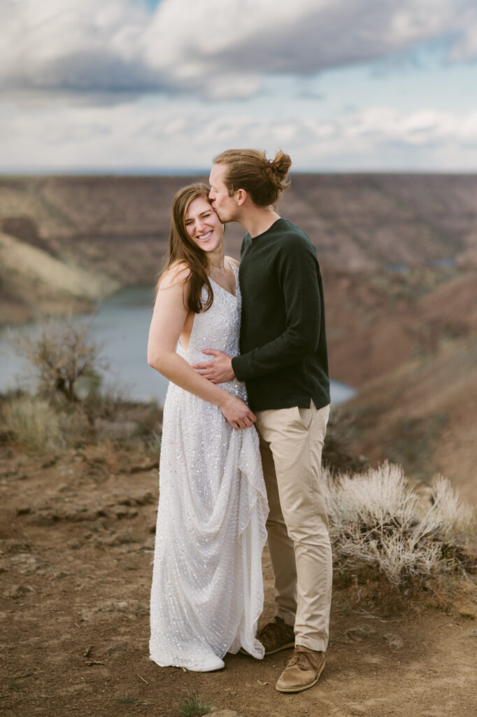 A man and woman stand at the Tam-a-lau Trail overlook at Cove Palisades State Park in Oregon. The woman is wearing a white dress and holding it slightly off the ground. The man has both hands around her waist and is kissing her temple as she smiles with her eyes closed.