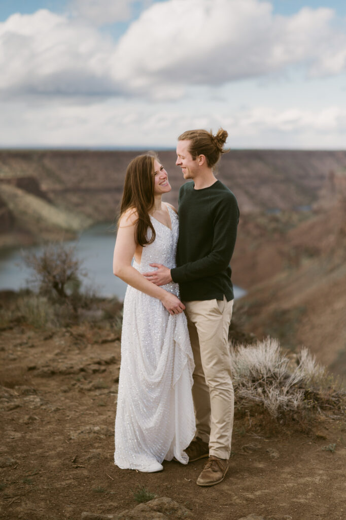 A man and woman stand at the Tam-a-lau Trail overlook at Cove Palisades State Park in Oregon. The woman is wearing a white dress and holding it slightly off the ground. The man has both hands around her waist and they are looking at one another, smiling.