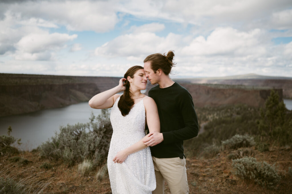 A man and woman standing on the Tam-a-lau Trail at Cove Palisades State Park. The woman is holding her dress with one hand and playing with her hair with her other hand. The man rests one hand on the woman's elbow and leans down to kiss her.