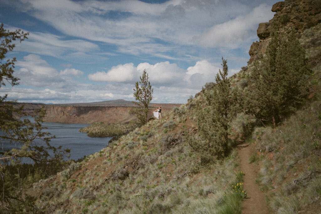 A pulled back photo showing a very small man and woman off in the distance on the Tam-a-lau Trail at Cove Palisades State Park in Oregon. The woman has thrown her white dress back so it flows out behind her.