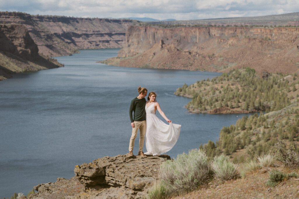 A man and woman stand on an overlook at the Tam-a-lau Trail at Cove Palisades State Park in Oregon. The woman has one arm around the man, the other is flicking the train of her dress out. She is looking down at it while the man looks at her.